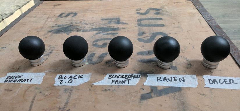 Bu görsel, çeşitli siyah boyaların matlığını karşılaştırmak amacıyla düzenlenmiş bir test gösterimini içermektedir. Soldan sağa doğru, "Dulux Rich Matt," "Black 2.0," "Blackboard Paint," "Raven," ve "Daler" gibi farklı marka ve türlerdeki siyah boyalar yer almaktadır. Vantablack, bu tür boyalarla kıyaslandığında, en düşük yansıma oranına sahip olup, ışığı en fazla absorbe etmesiyle dikkat çeker.